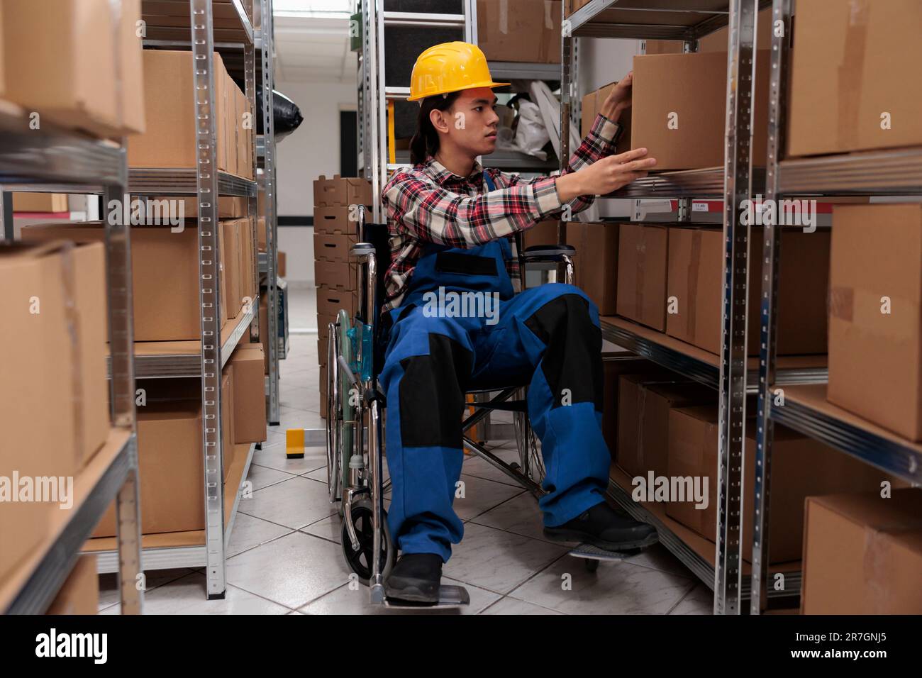 Jeune ouvrier d'entrepôt asiatique en fauteuil roulant prenant des colis de l'étagère. Préparateur de commande de magasin avec incapacité physique à rechercher une boîte en carton pendant le travail dans la salle de stockage Banque D'Images