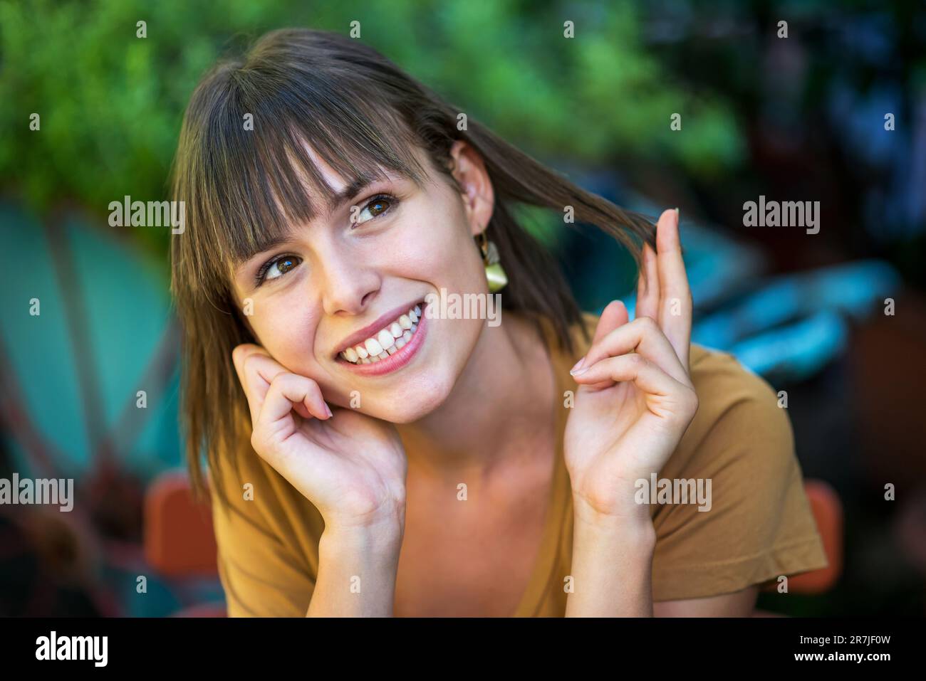 Portrait d'une jeune femme positive avec un sourire ondoyant, en regardant loin avec la main au menton et en touchant les cheveux tout en étant assise sur une chaise contre un arbre vert flou Banque D'Images