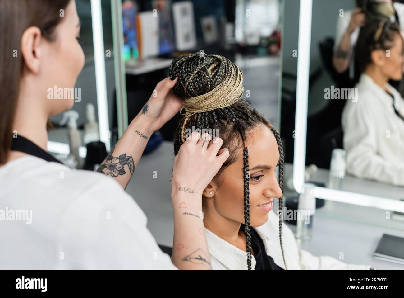 salon de coiffure, coiffeur tatoué faisant un pain de cheveux à la cliente féminine avec des braïdes, des femmes gaies, la satisfaction de la clientèle, la cliente dans le salon de beauté, le préposé à la beauté Banque D'Images