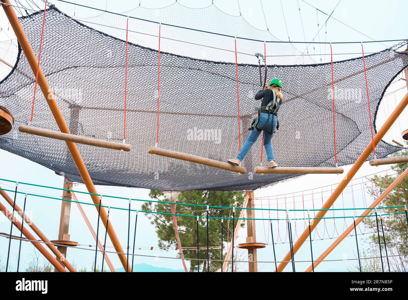 Adolescente dans l'équipement de harnais d'escalade, casque de sécurité vert de sport. Parc d'attractions de corde. Fixation fixant le mousqueton à la corde de sécurité. Hangin Banque D'Images