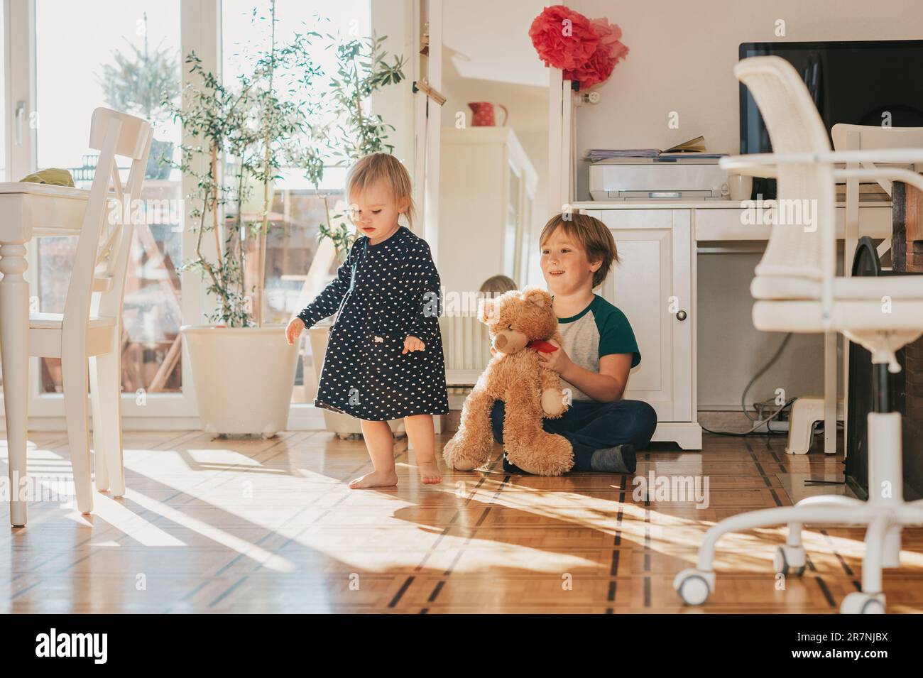 Deux enfants amusants jouant avec l'ours en peluche à la maison dans la salle de séjour blanche remplie de lumière du soleil Banque D'Images