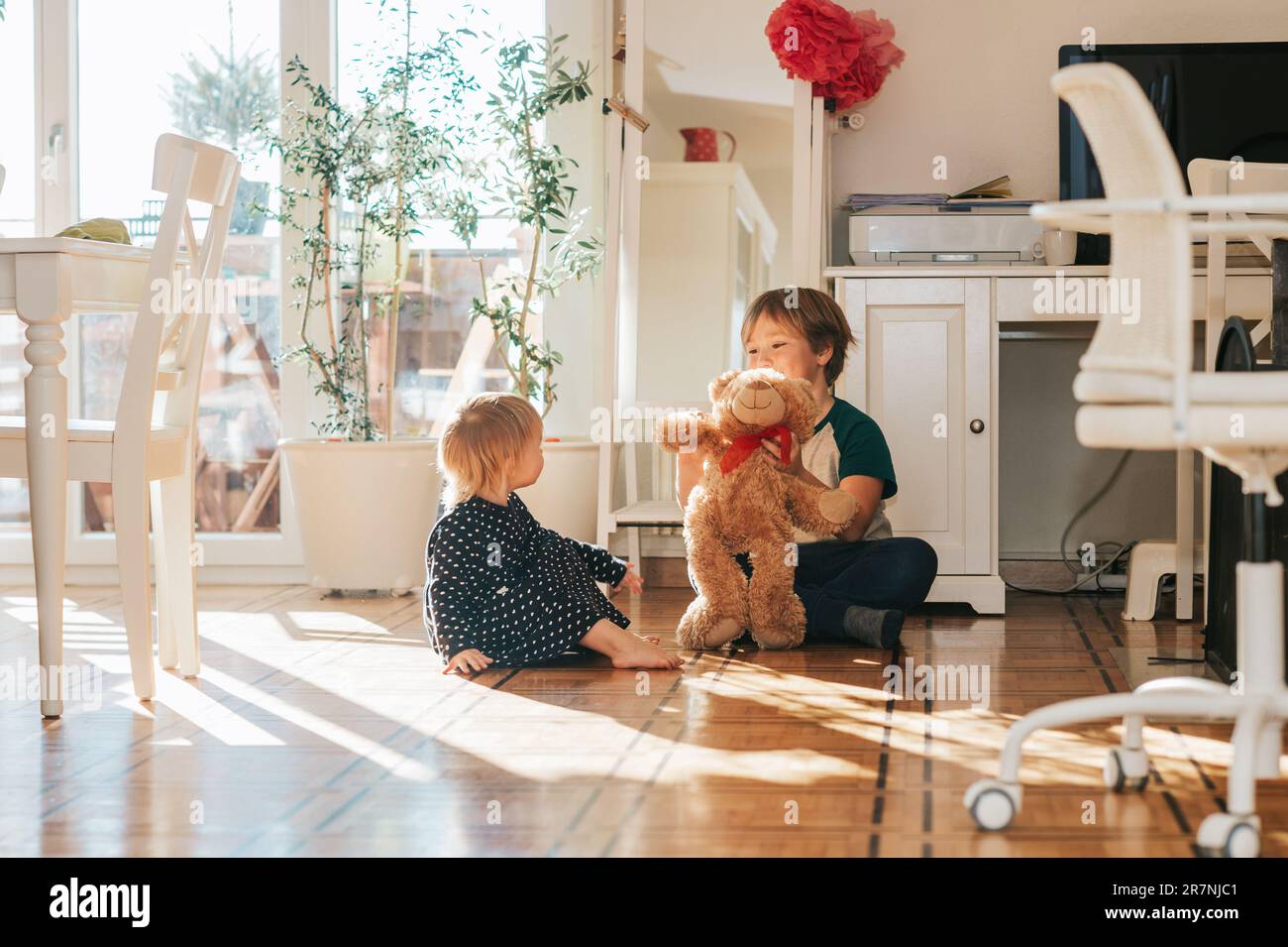 Deux enfants amusants jouant avec l'ours en peluche à la maison dans la salle de séjour blanche remplie de lumière du soleil Banque D'Images