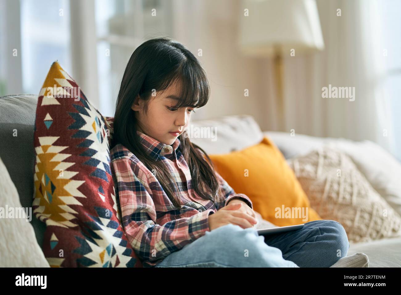 petite fille asiatique de sept ans assise sur un canapé de famille à la maison à l'aide d'un ordinateur tablette numérique Banque D'Images