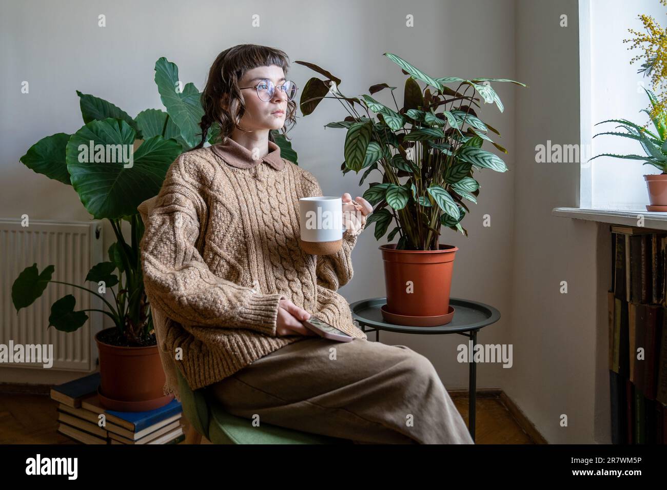 Jeune fille pensive du millénaire assise avec téléphone et tasse de thé dans la jungle urbaine maison donnant sur la fenêtre Banque D'Images