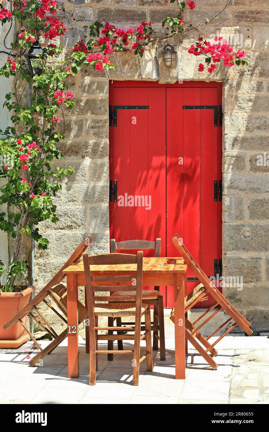 Belle vue sur une allée dans la ville de Lefkada, avec une table en bois et 4 chais en bois autour, une porte en bois rouge d'une maison en pierre et des plantes. Banque D'Images