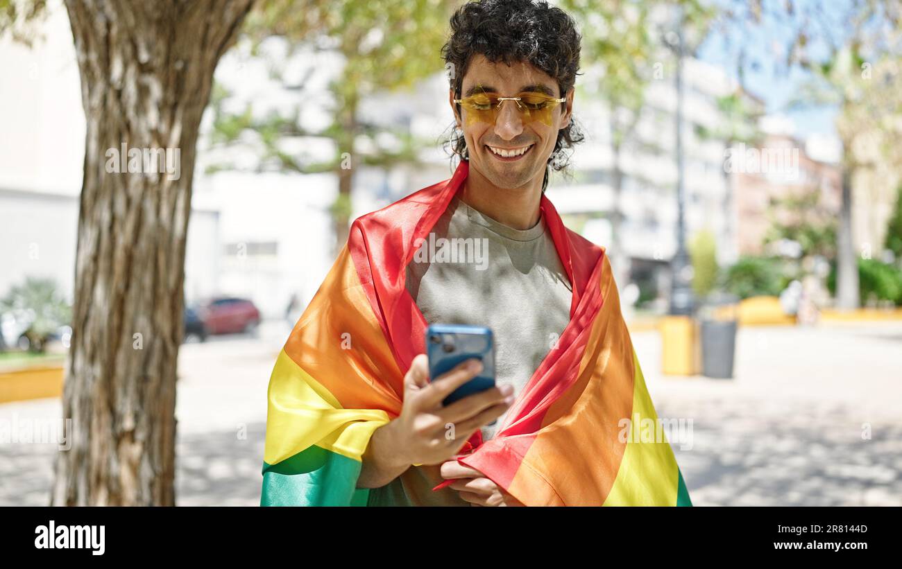 Jeune homme hispanique portant le drapeau arc-en-ciel à l'aide d'un smartphone au parc Banque D'Images