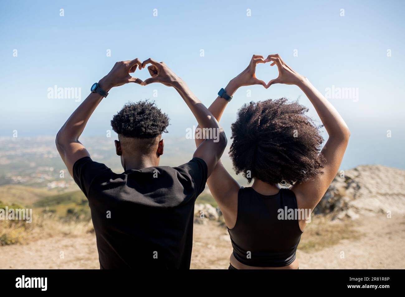 Jeune couple sportif noir profitant de l'entraînement à l'extérieur faisant le coeur avec des mains signe, debout sur des rochers à l'océan, vue arrière Banque D'Images