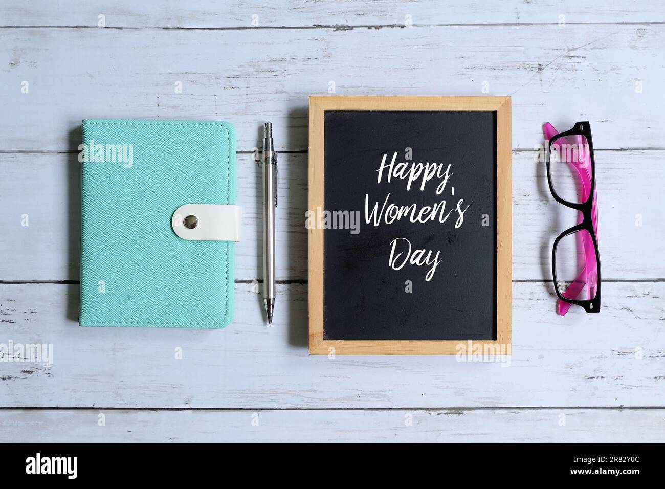 Vue de dessus du tableau noir écrit « HAPPY WOMEN'S DAY » avec un stylo pour ordinateur portable et des lunettes de soleil sur fond blanc en bois. Banque D'Images