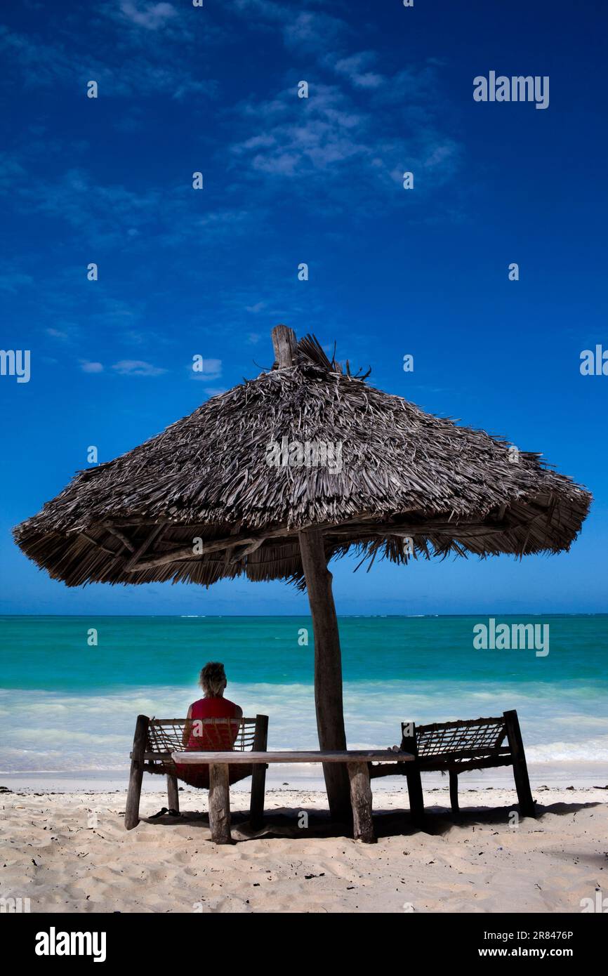 Un touriste cherche un abri du soleil sous un parapluie de plage fait de feuilles de plam en Tanzanie. Banque D'Images