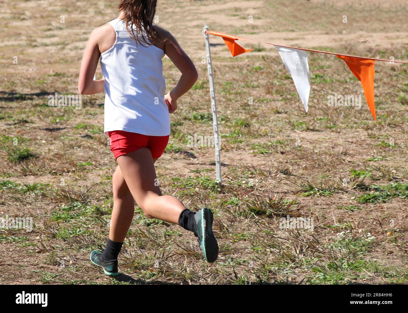 Vue arrière d'une lycéenne qui court dans une course de cross-country sur l'herbe et la terre à côté des drapeaux colorés marquant le parcours portant un haut blanc et RE Banque D'Images