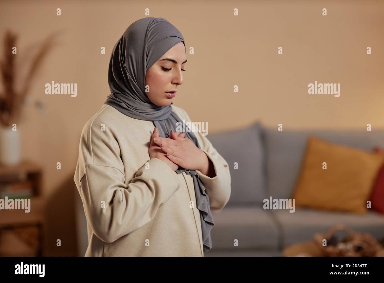 Jeune femme islamique en hijab gris et sweatshirt beige exprimant sa gratitude à son Dieu pendant la prière tout en gardant ses mains sur la poitrine Banque D'Images