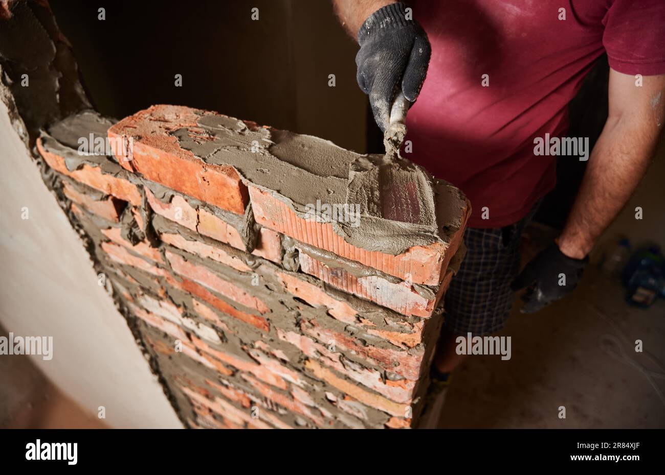 Gros plan des mains masculines dans des gants de travail portant des briques dans un bâtiment en construction. Homme bricklayer constructeur application de mortier de ciment sur les briques avec l'outil de truelle. Concept de construction en maçonnerie. Banque D'Images