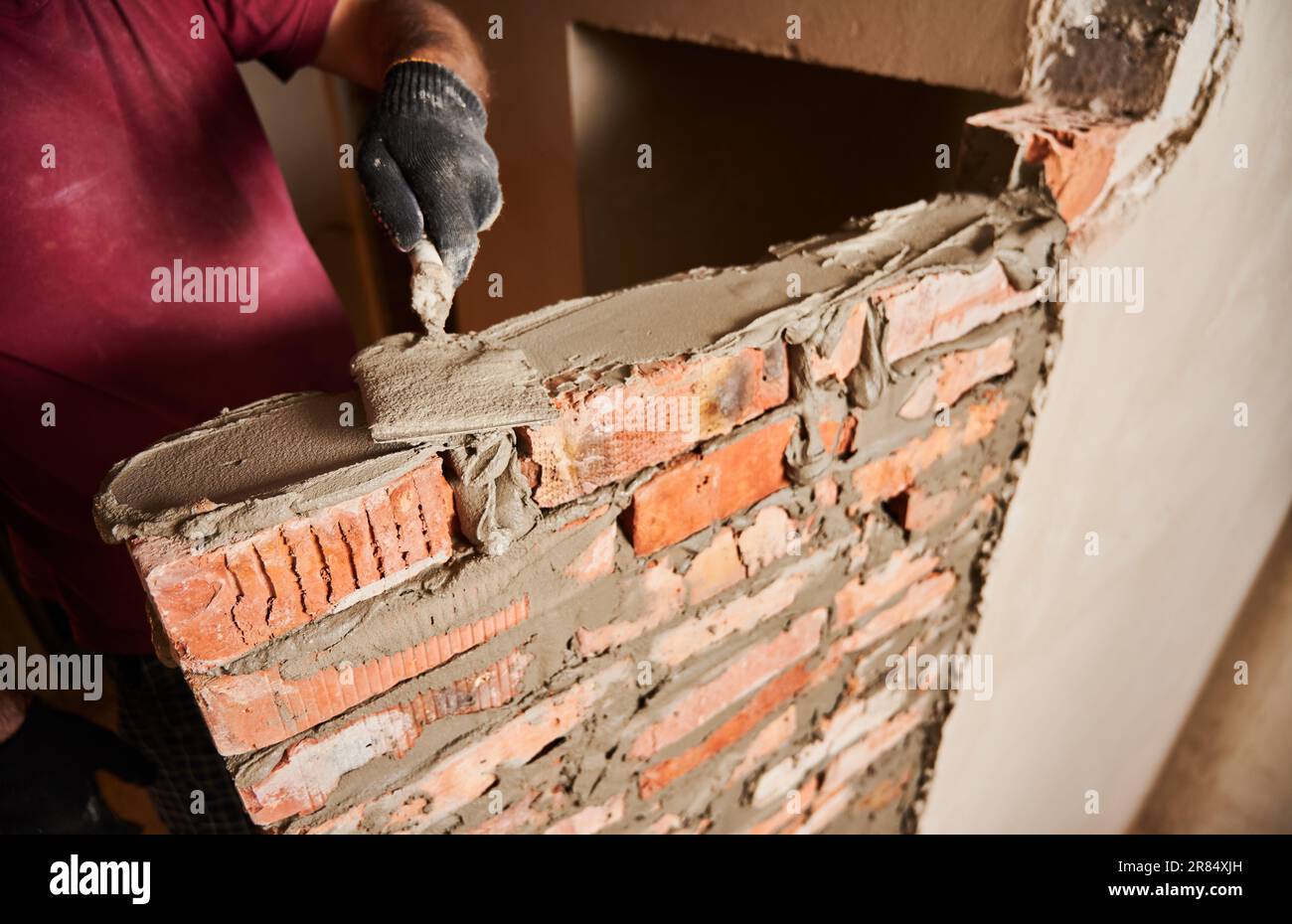 Gros plan de l'homme main dans le gant de travail pose de briques dans le bâtiment en construction. Travailleur masculin appliquant du mortier de ciment sur des briques avec un outil à truelle. Concept de construction en maçonnerie. Banque D'Images