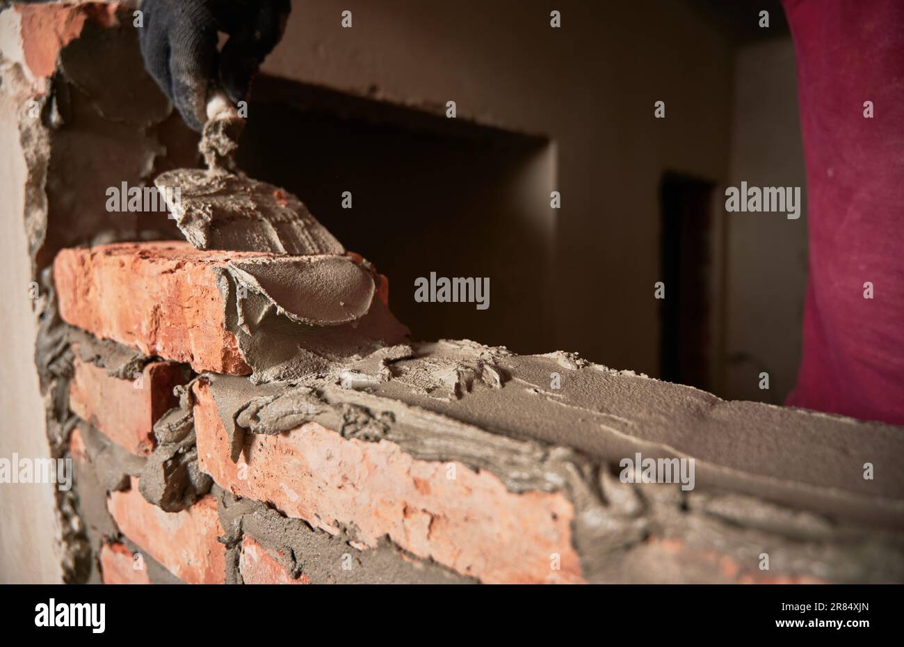 Gros plan de l'homme main dans le gant de travail pose de briques dans le bâtiment en construction. Travailleur masculin appliquant du mortier de ciment sur des briques avec un outil à truelle. Concept de construction en maçonnerie. Banque D'Images