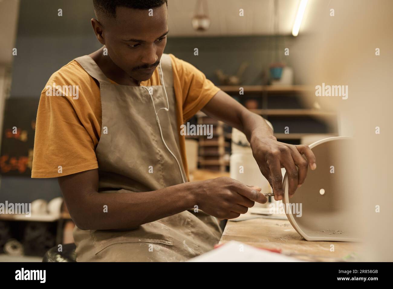 Un jeune céramiste africain fait des trous dans un pot à un banc dans un atelier Banque D'Images