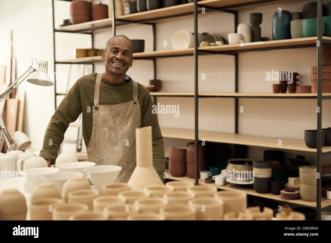 Céramiste africain souriant debout près d'un banc et des étagères pleines de pots Banque D'Images
