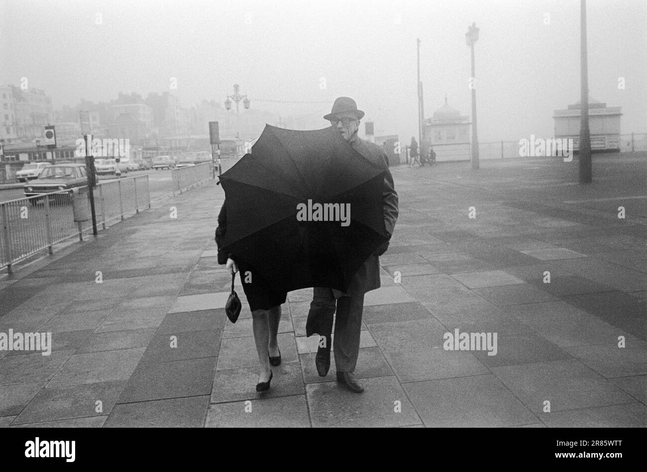 Angleterre mauvais temps pluvieux. Couple d'âge moyen marchant pendant le bord de mer anglais traditionnel mauvais temps promenade de front de mer Brighton. Brighton, Sussex, Grande-Bretagne années 1969 1960 Royaume-Uni HOMER SYKES Banque D'Images