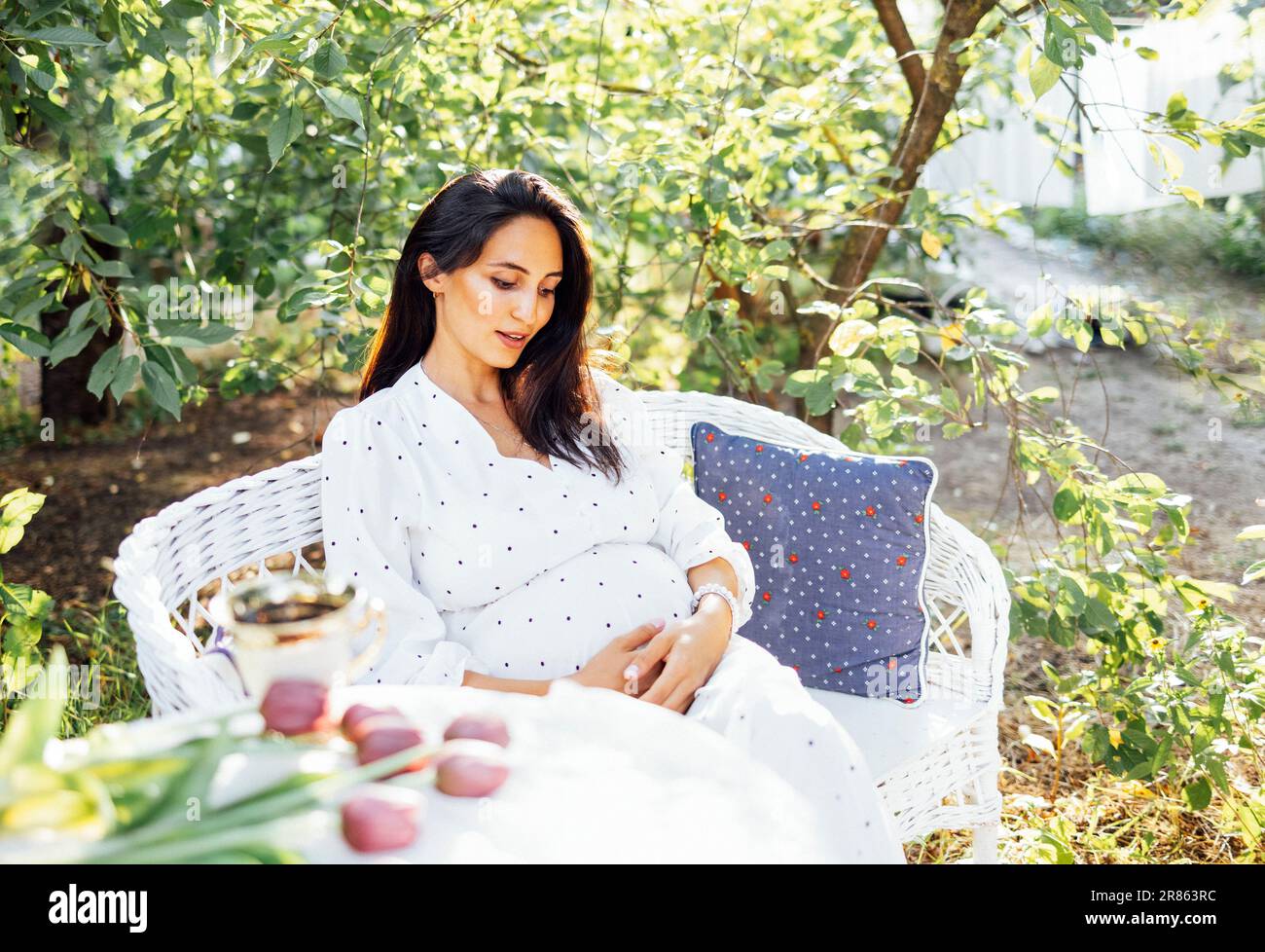 Une charmante brunette enceinte dans une élégante robe est assise sur un canapé en osier blanc avec oreillers à l'extérieur dans le jardin. La jeune femme attend bébé tient doucement h Banque D'Images