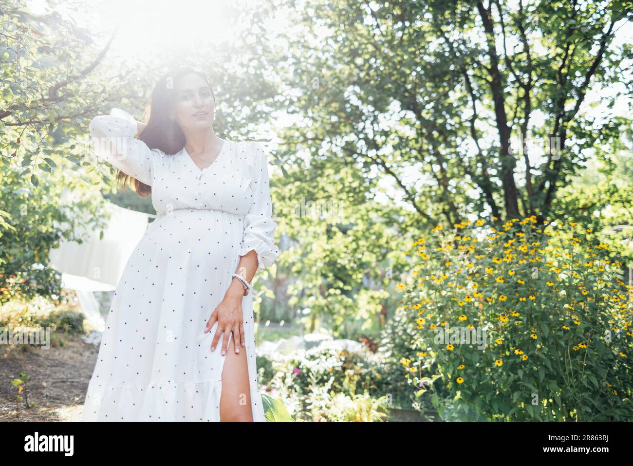 Charmante brunette enceinte en élégante robe blanche est debout à l'extérieur dans le jardin parmi les arbres et les fleurs. Mettez en évidence les rayons ensoleillés. Une jeune femme s'attend à Banque D'Images