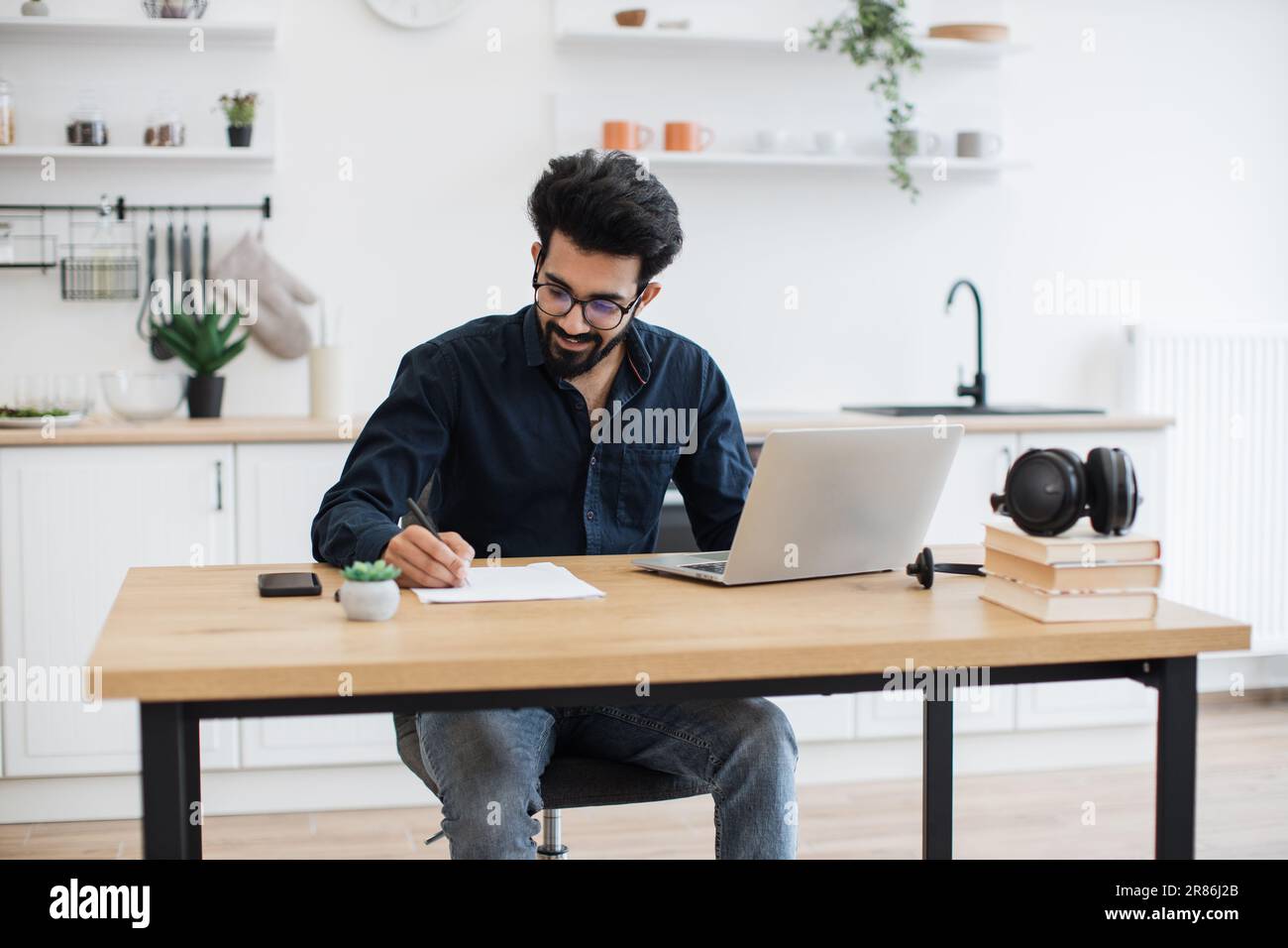 Beau indien portant une chemise sombre et des lunettes écrivant sur le document tout en restant à la maison pendant la mi-journée. Propriétaire d'entreprise efficace améliorant le contrat tout en vérifiant les données sur un ordinateur portable. Banque D'Images