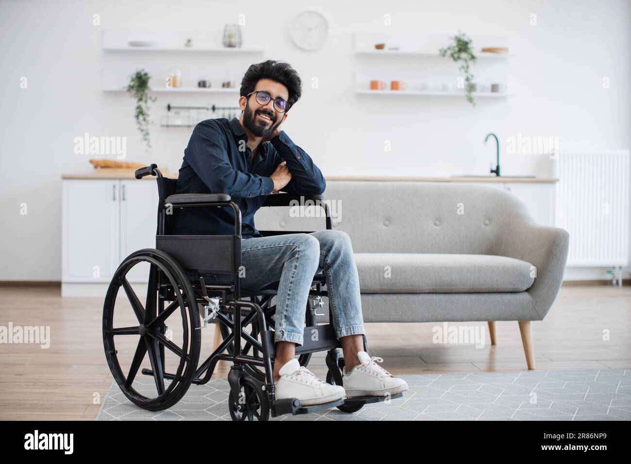 Portrait complet d'un utilisateur de fauteuil roulant arabe souriant à  l'appareil photo tout en se reposant dans la cuisine ouverte de  l'appartement moderne. Joyeux jeune homme dans des vêtements et des lunettes