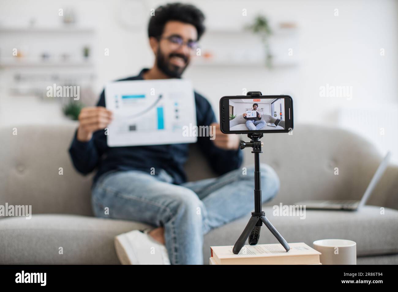 Un homme indien charmant dans des vêtements décontractés assis sur un canapé avec un graphique de rapport financier entre les mains tout en enregistrant la vidéo sur un smartphone moderne. Un financier qui a réussi à parler de statistiques sur la caméra. Banque D'Images