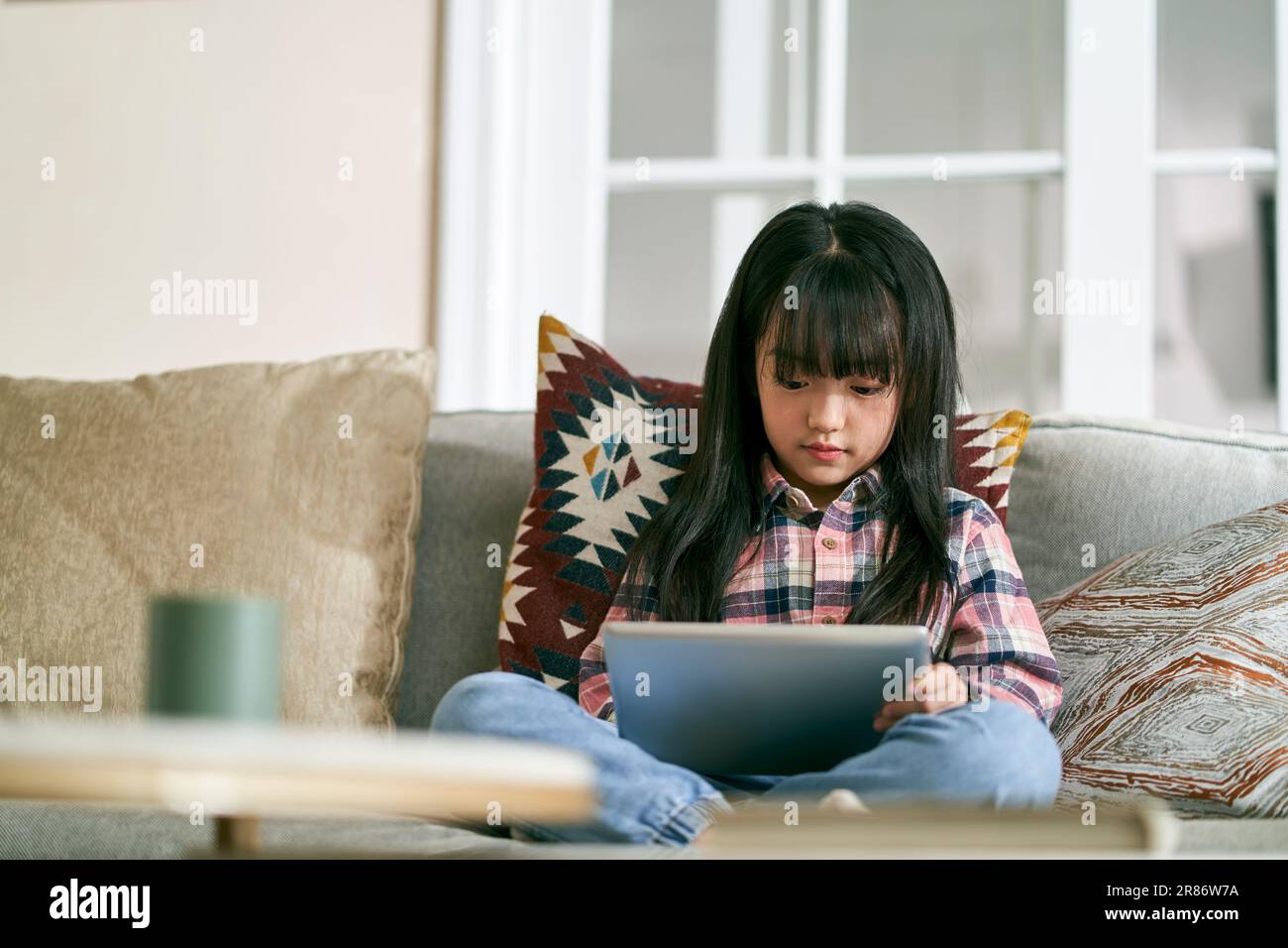 petite fille asiatique de sept ans assise sur un canapé de famille à la maison à l'aide d'un ordinateur tablette numérique Banque D'Images