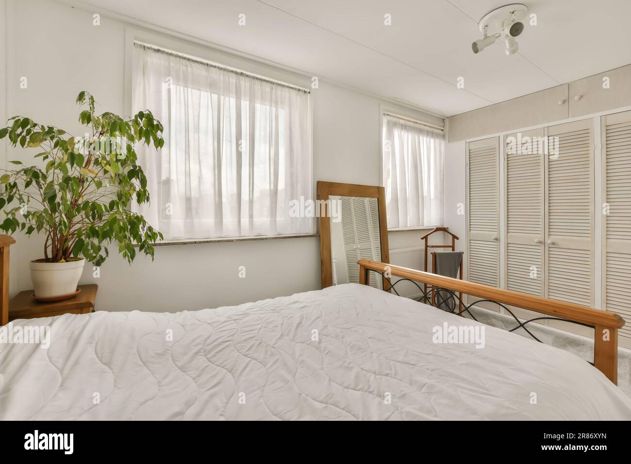 un lit dans une chambre avec des draps blancs et des rideaux sur la fenêtre derrière elle est une plante qui ressemble à un arbre Banque D'Images
