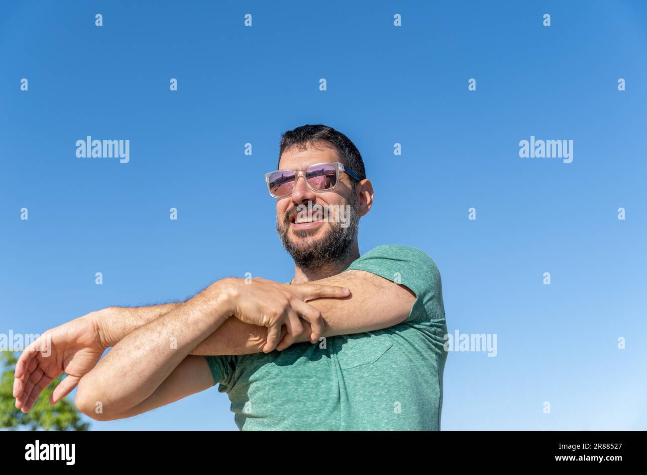 Vue de face d'un bel homme barbu avec des lunettes de soleil souriant et étirant ses muscles dans un champ avec un ciel bleu en arrière-plan Banque D'Images