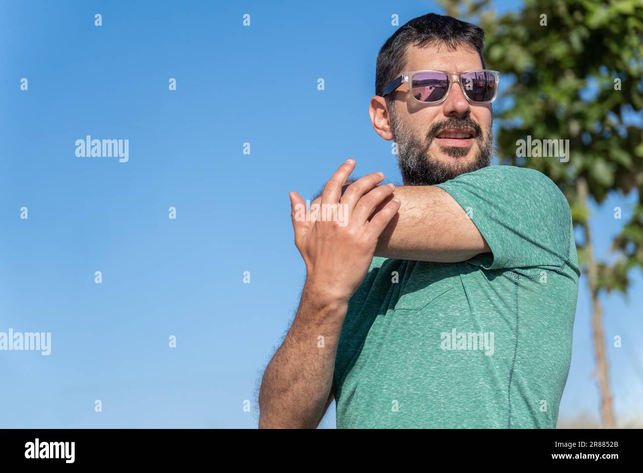 Vue avant d'un bel homme barbu avec des lunettes de soleil qui s'étendent sur ses épaules dans un champ avec un ciel bleu en arrière-plan Banque D'Images