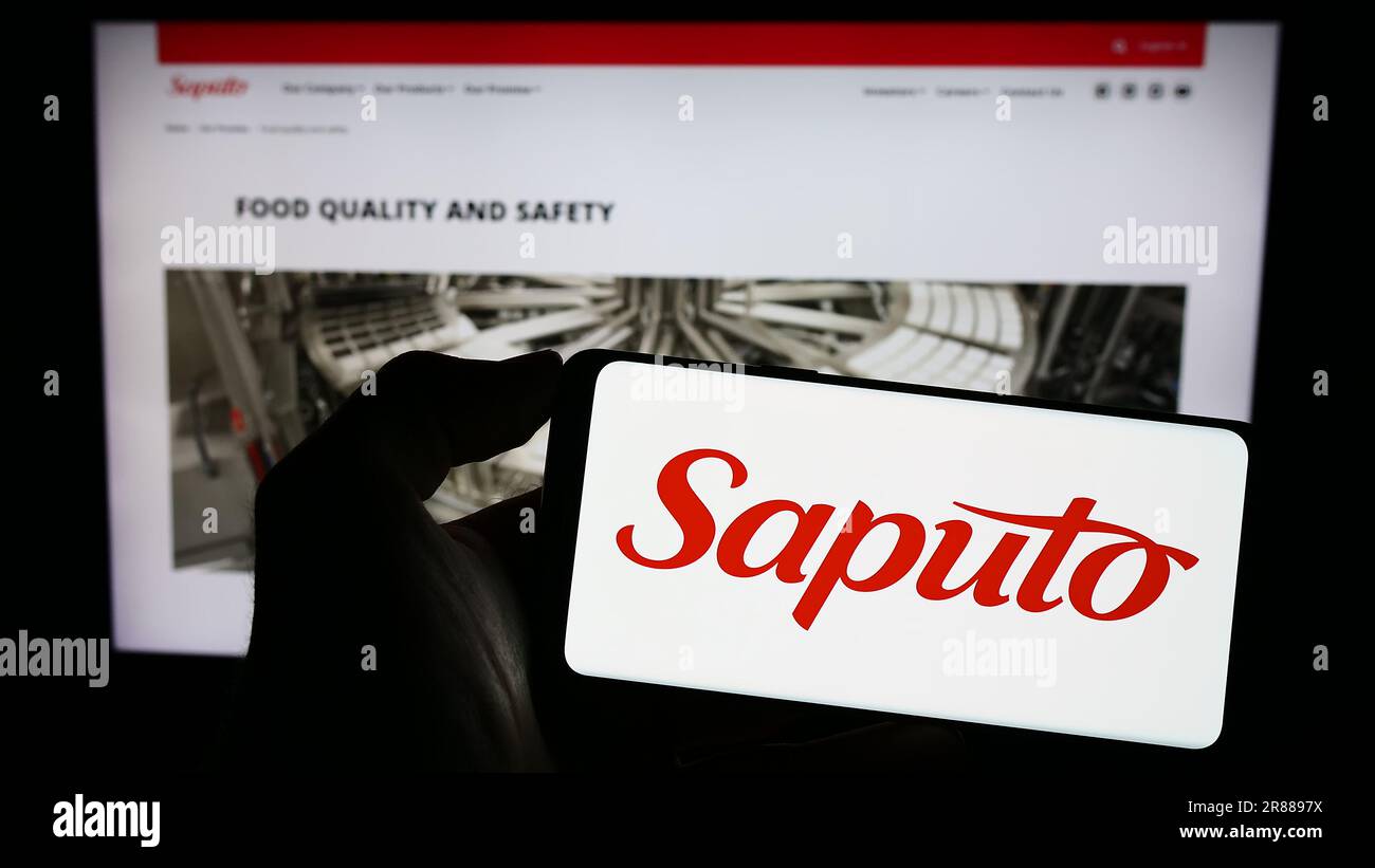 Personne tenant un téléphone cellulaire avec le logo de la société laitière canadienne Saputo Inc. À l'écran devant la page Web des affaires. Mise au point sur l'affichage du téléphone. Banque D'Images