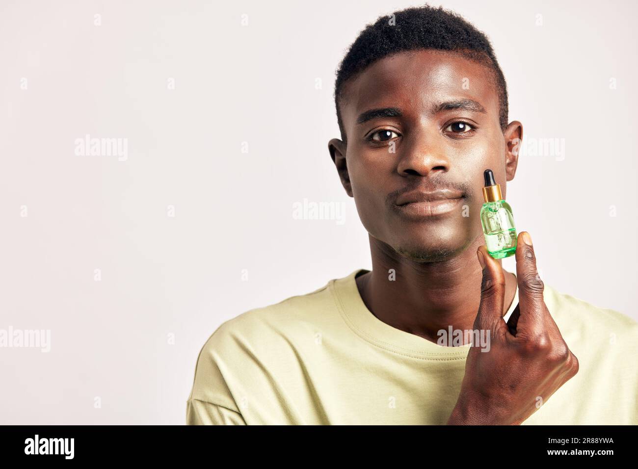 Un homme africain dans un t-shirt avec une petite bouteille verte de lotion pour la peau. Homme noir en studio avec la beauté, la santé et la procédure naturelle de peau. Bien-être, cosme Banque D'Images
