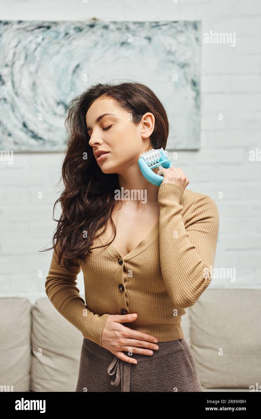 Jeune brunette et femme détendue en pull brun massant les nœuds lymphatiques avec masseur avec poignée tout en se tenant dans la maison floue, rituel d'auto-soin et Banque D'Images