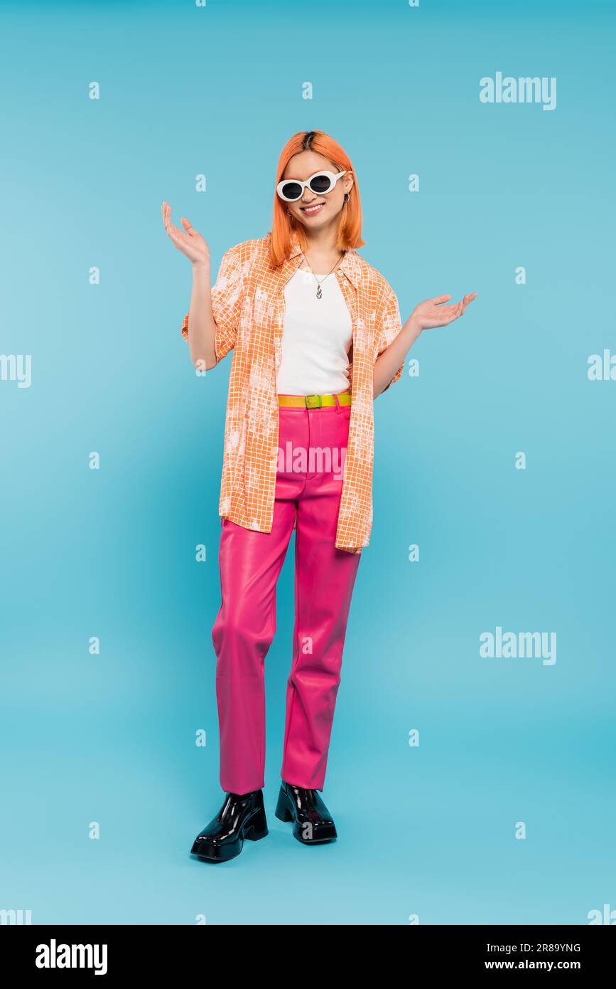 style personnel, femme asiatique heureuse avec des cheveux teints debout dans une tenue décontractée et des lunettes de soleil, gesturant avec les mains sur fond bleu vif, shi orange Banque D'Images
