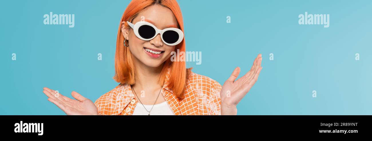 positive, jeune femme asiatique avec des cheveux teints debout dans une tenue décontractée et des lunettes de soleil, gestante avec les mains sur fond bleu vif, chemise orange, ne Banque D'Images