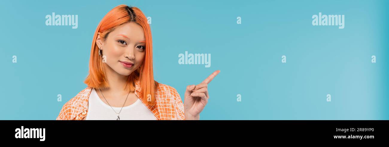 démonstration, jeune femme asiatique aux cheveux rouges teints regardant la caméra sur fond bleu vif, chemise orange, pointant avec le doigt, montrant quelque chose de mince Banque D'Images
