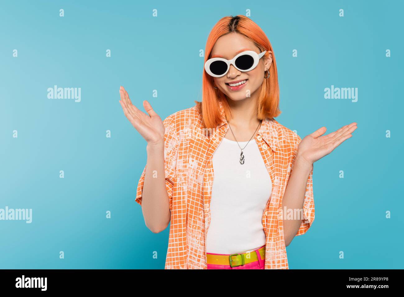 bonheur, jeune femme asiatique avec des cheveux teints debout dans une tenue décontractée et des lunettes de soleil, gesturant avec les mains sur fond bleu vif, chemise orange, n Banque D'Images