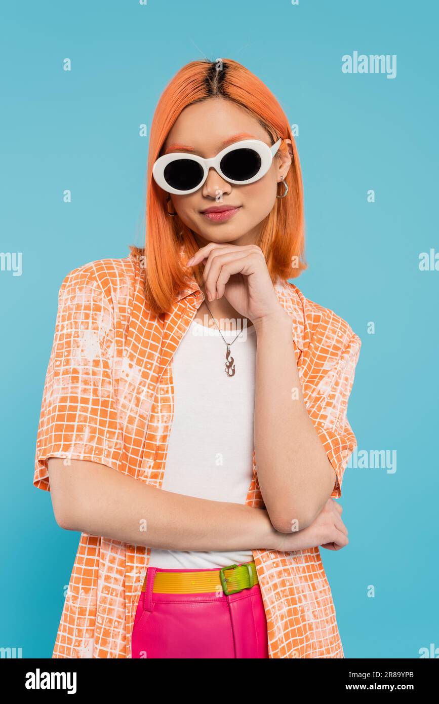 generation z, jeune femme asiatique avec des cheveux teints debout dans une tenue décontractée et des lunettes de soleil, regardant l'appareil photo sur fond bleu vif, chemise orange, r Banque D'Images