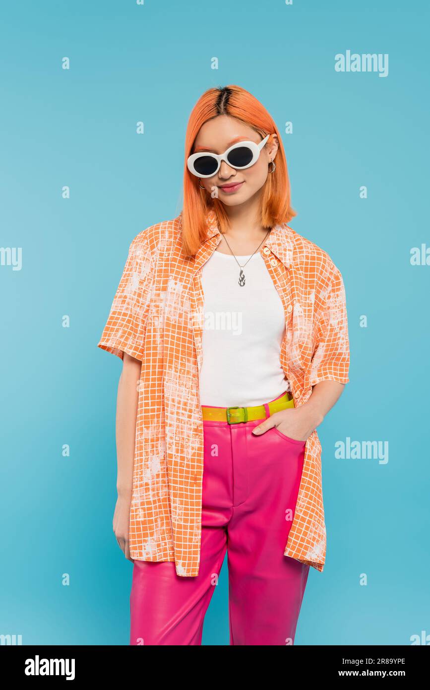 tenue décontractée, femme asiatique souriante avec cheveux teints et lunettes de soleil debout avec main dans la poche sur fond bleu vif, chemise orange, cheveux rouges, mode Banque D'Images