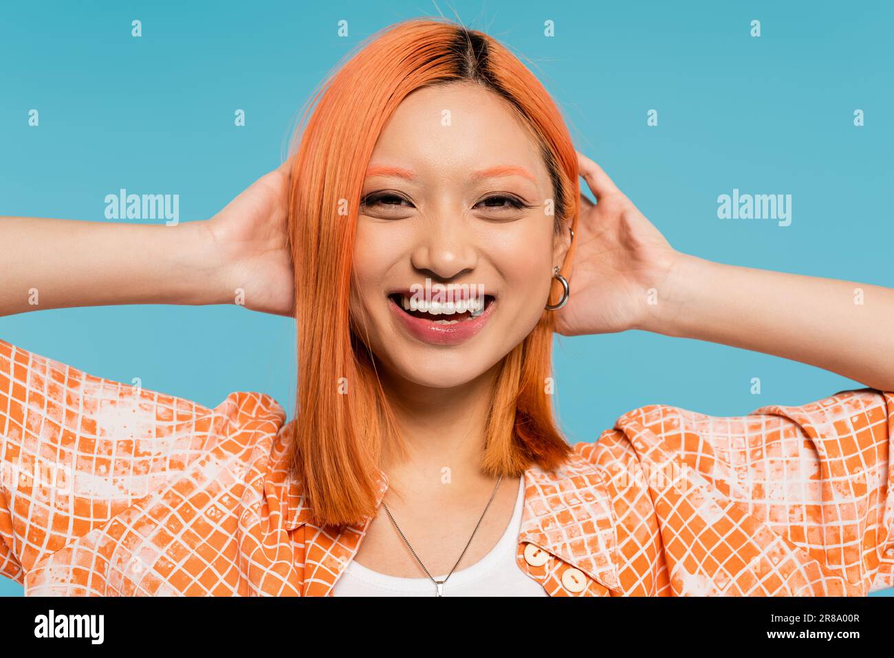 positivité, sourire éclatant, jeune femme asiatique aux cheveux teints debout dans une chemise orange et souriant sur fond bleu, tenue décontractée, liberté, gai Banque D'Images