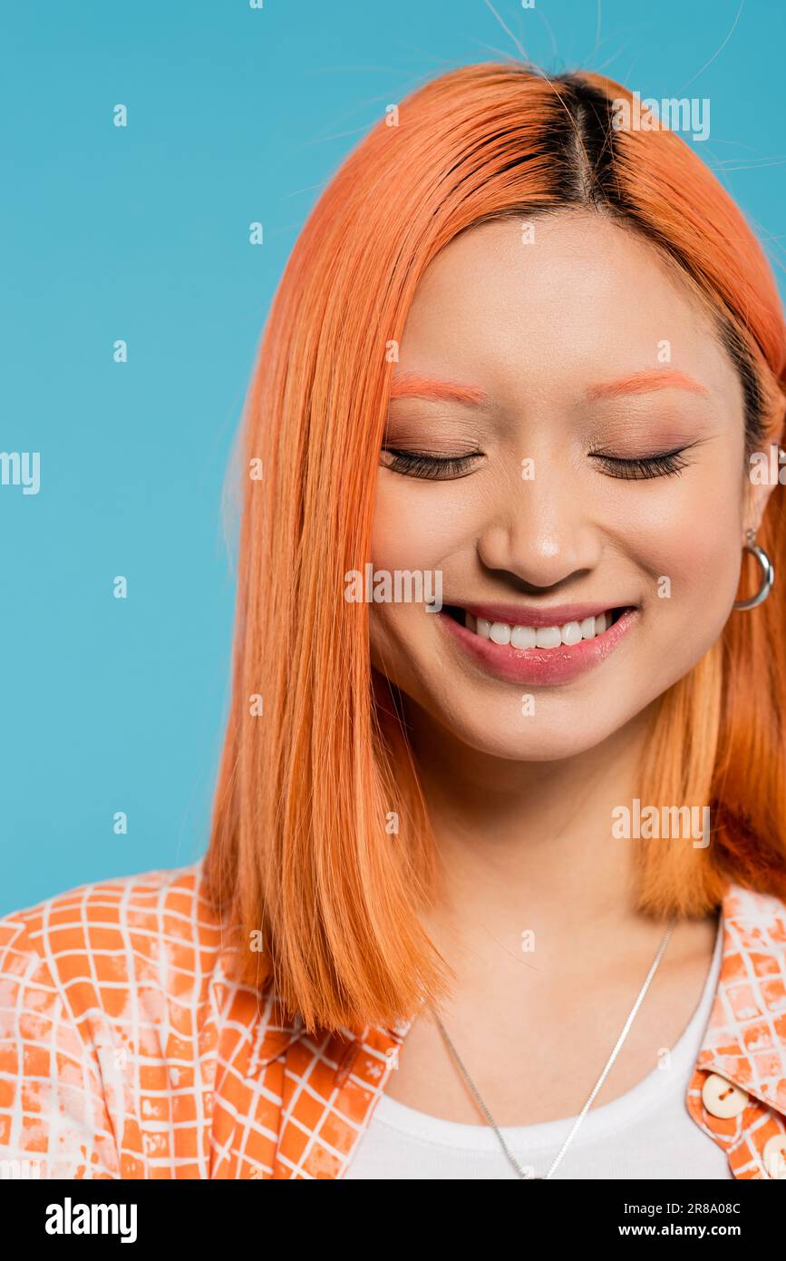 portrait, visage gai, jeune femme asiatique avec cheveux courts et teints, maquillage naturel et boucles d'oreilles en forme de cerceau souriant sur fond bleu, chemise orange, genres Banque D'Images