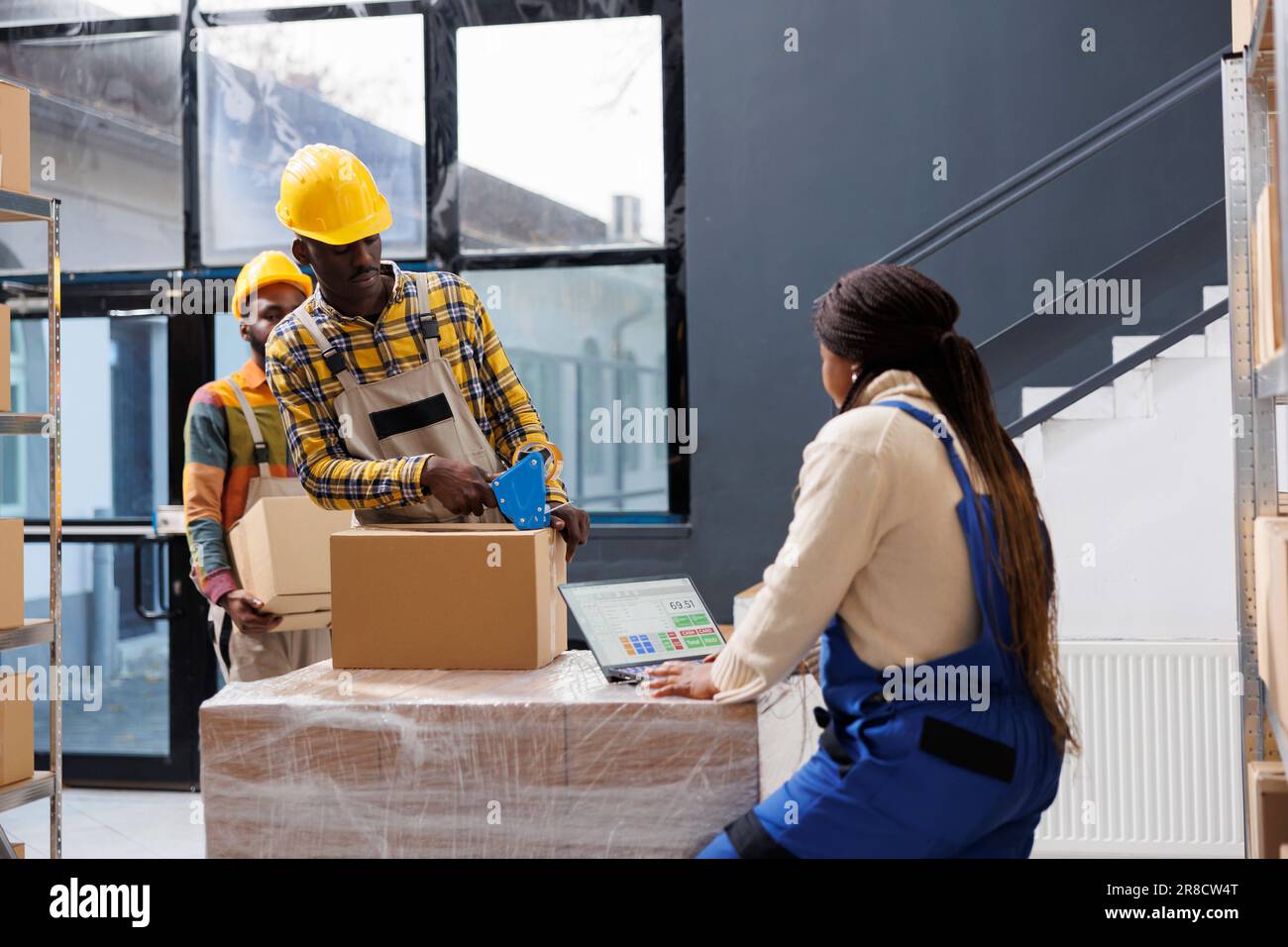 Hommes et femmes afro-américains préparateurs de commande travaillant dans un entrepôt de détail. Le gestionnaire d'emballage emballant la boîte en carton avec le distributeur de ruban adhésif pendant que le responsable du magasin vérifie le bon de cueillette sur l'ordinateur portable Banque D'Images
