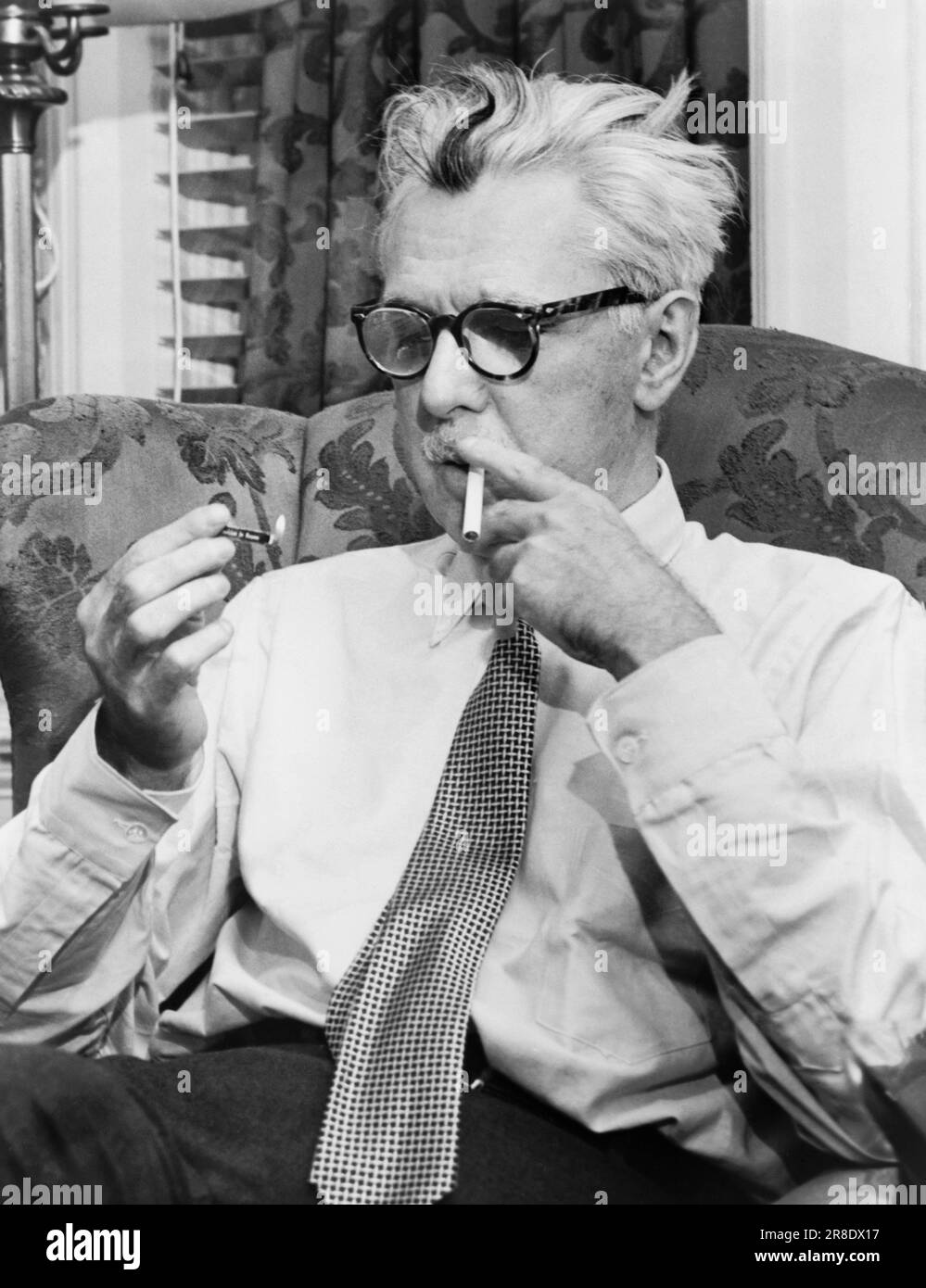 New York, New York: 1954 auteur, caricaturiste et humoriste James Thurber assis dans une chaise éclairant une cigarette. Banque D'Images
