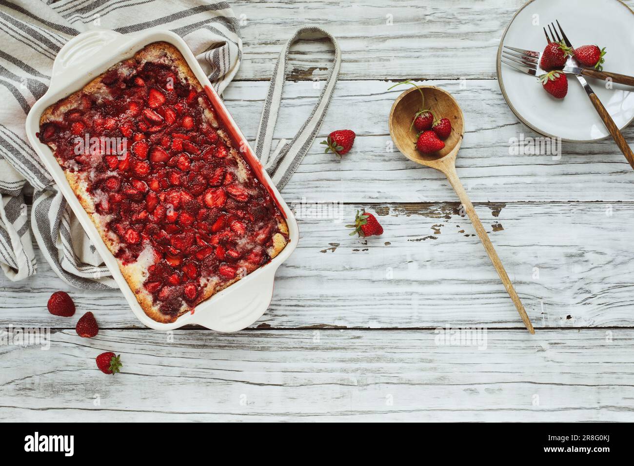 Vue de dessus à angle élevé de cordonnier de fraise maison sucrée ou Sonker cuit dans une poêle en céramique rouge avec tablier et cuillère en bois sur une table rustique. Banque D'Images