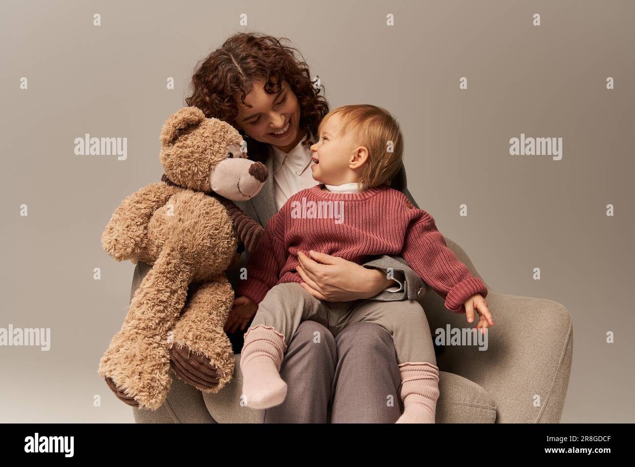 qualité du temps en famille, parent et carrière, femme d'affaires heureuse tenant l'ours en peluche près de la petite fille sur fond gris, assis sur un fauteuil, travail Banque D'Images