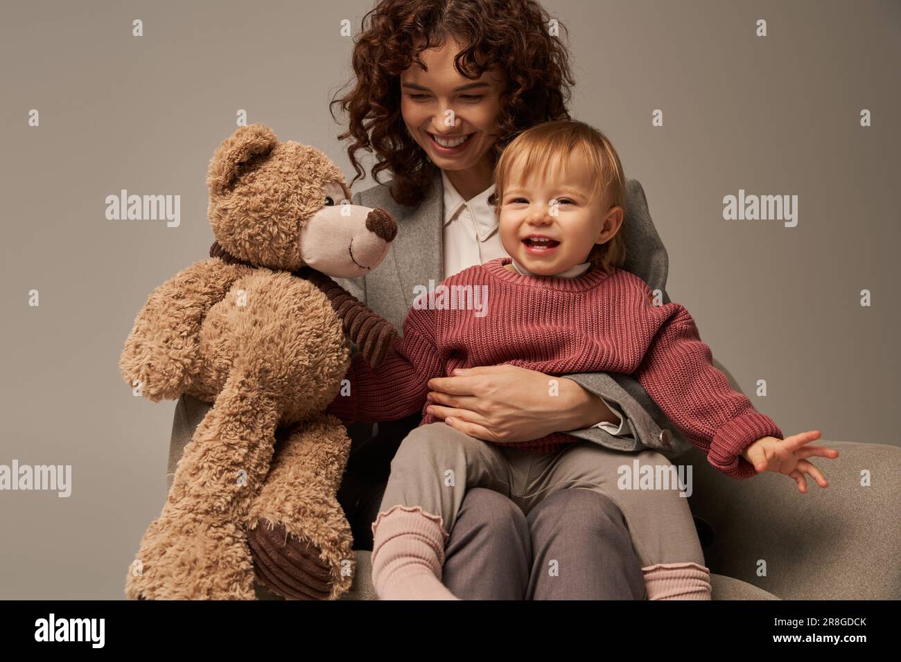 qualité du temps de famille, parent et carrière, gaie femme d'affaires tenant l'ours en peluche près de la petite fille sur fond gris, assis sur un fauteuil, wo Banque D'Images