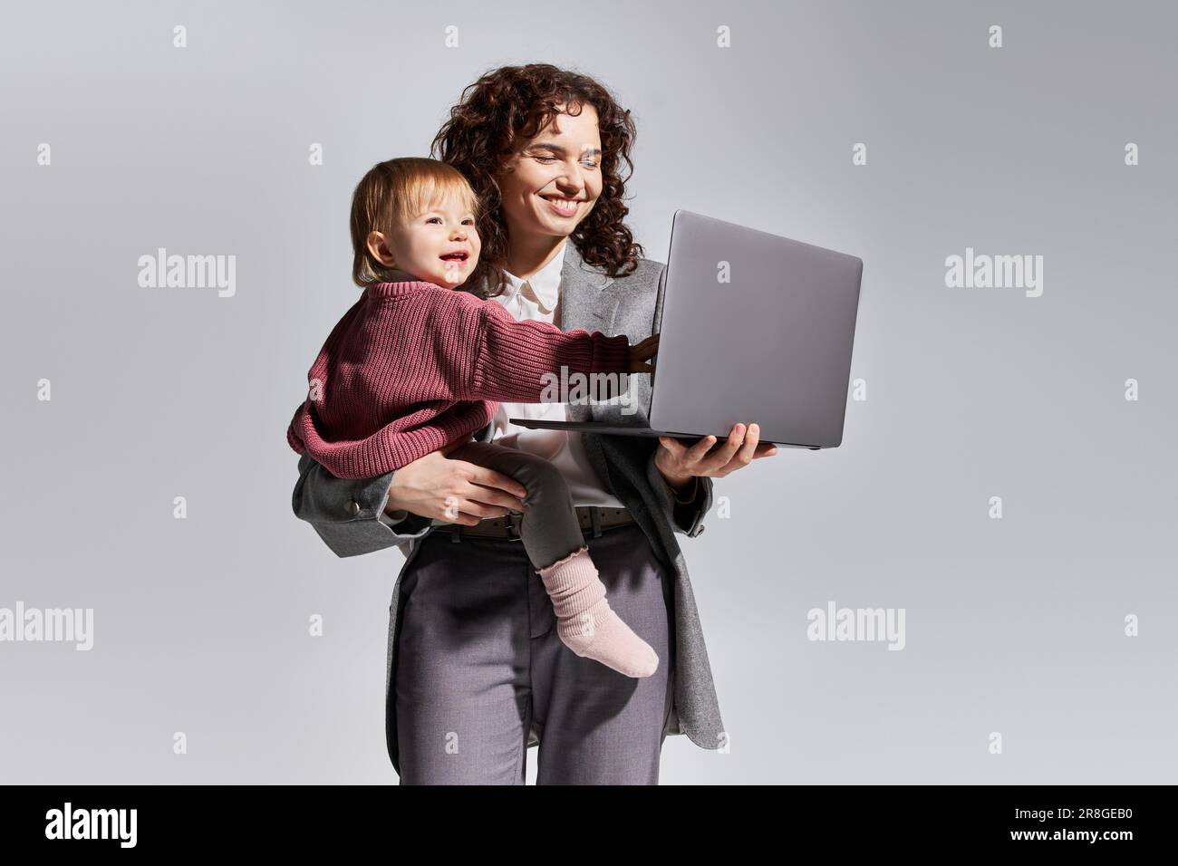 gestion du temps, mère travaillant tenant un ordinateur portable et petite fille dans les bras sur fond gris, l'harmonie de la vie de travail, carrière et famille, parent moderne Banque D'Images