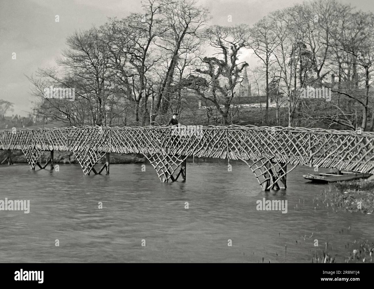 Une passerelle au-dessus d'une rivière au Royaume-Uni à l'époque victorienne. L'utilisation de branches d'arbre rustiques tissées décoratives pour la main courante et son côté donnent à la traversée un aspect très distinct et inhabituel. Une femme pose sur le pont et un punt est attaché contre la rive droite - une photographie victorienne vintage. Banque D'Images