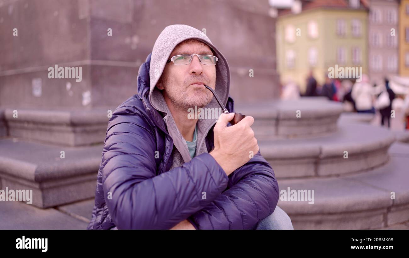 Portrait d'un homme adulte avec des lunettes assis dans la capuche sur la place et fumant un pipe de tabac sur la place du Palais, dans la vieille ville de Varsovie Banque D'Images