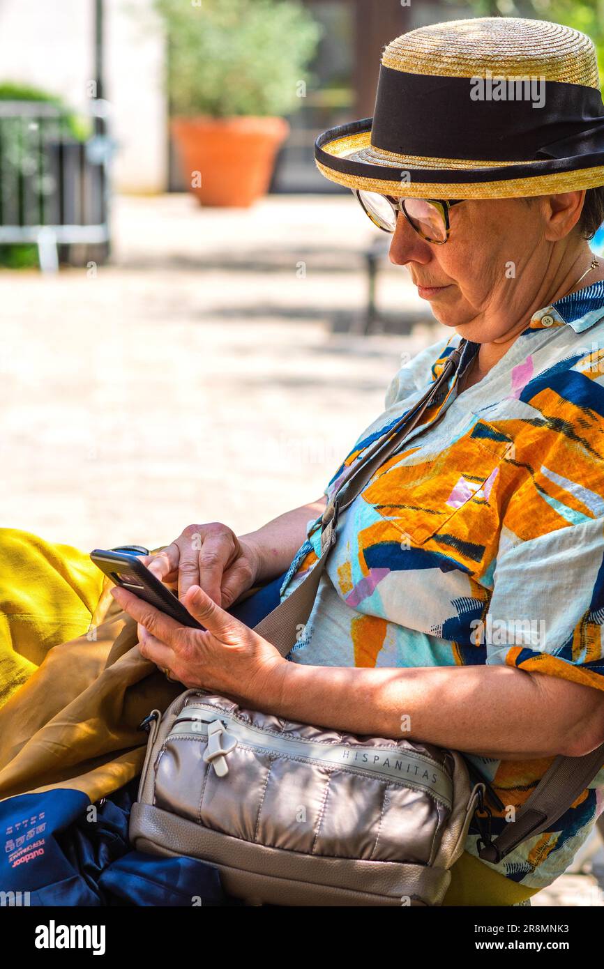 Femme mature dans chapeau de paille en centre ville banc jardin tapotant un message sur téléphone mobile - Tours, Indre-et-Loire (37), France. Banque D'Images